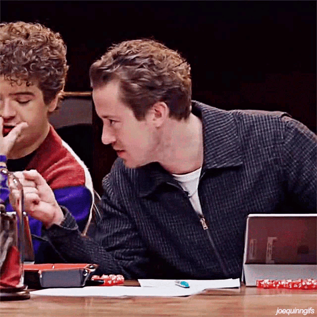 two men are sitting at a table talking to each other and one of them is holding a bottle .