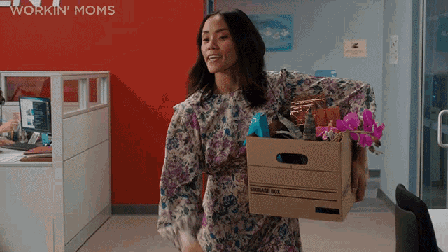 a woman is carrying a cardboard box in front of a wall that says workin moms