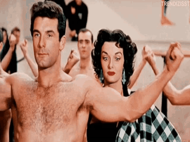 a man without a shirt is standing next to a woman in a plaid dress who is holding his arm .
