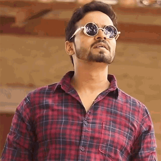 a man wearing round sunglasses and a plaid shirt looks up