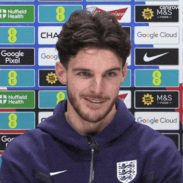a man wearing a purple hoodie stands in front of a wall of advertisements including google pixel and m & s food