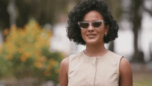 a woman wearing sunglasses and a tan dress smiles for the camera