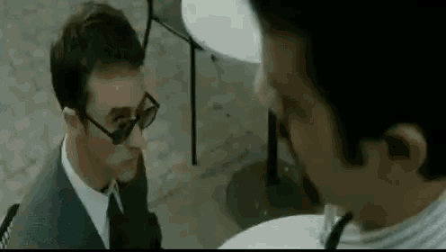 a man in a suit and tie is sitting at a table with another man wearing sunglasses .