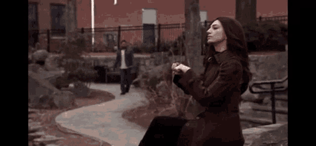 a woman in a brown coat is sitting on a bench in a park
