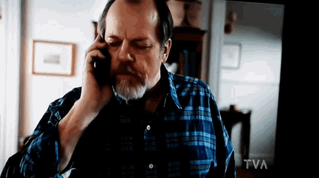 a man in a blue plaid shirt is talking on a cell phone in front of a tv screen that says tva