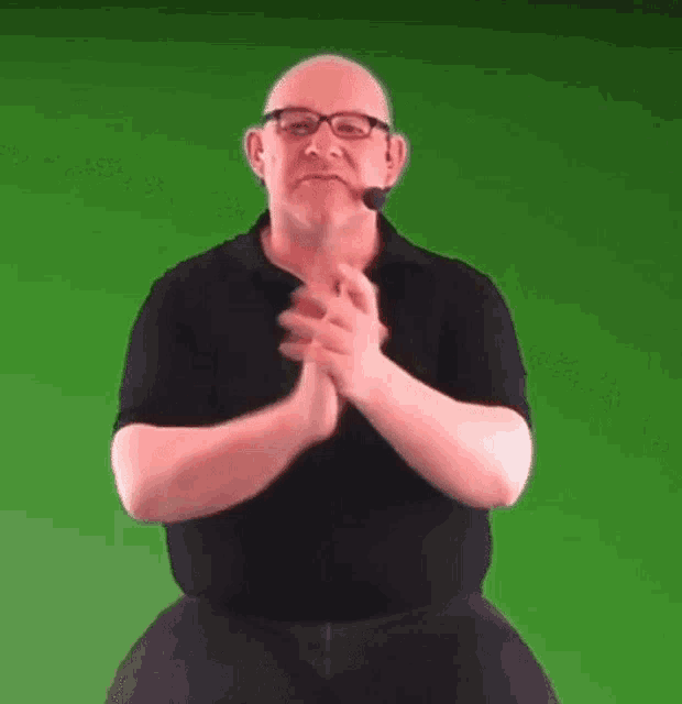 a man wearing glasses and a microphone is clapping his hands on a green screen .