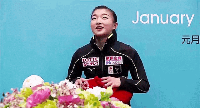 a woman in a black jacket is standing in front of a table with flowers and a blue background .