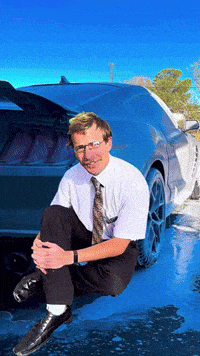 a man in a white shirt and tie sits on the ground in front of a car