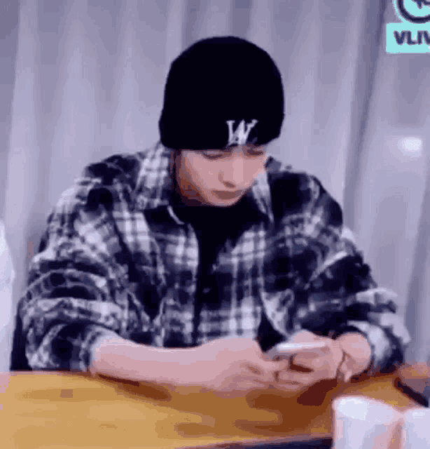 a young man wearing a plaid shirt and a black beanie is sitting at a table looking at his phone .