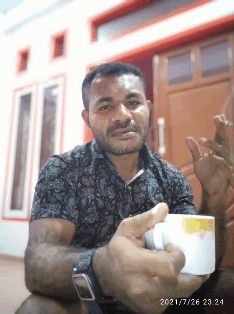 a man in a black shirt is holding a cup of coffee in front of a building with the date 2021/7/26 23:24