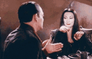 a man and a woman are sitting at a table talking to each other . the woman is wearing a black dress .