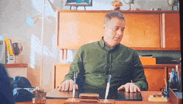 a man in a green shirt is sitting at a desk with two pens in front of him .