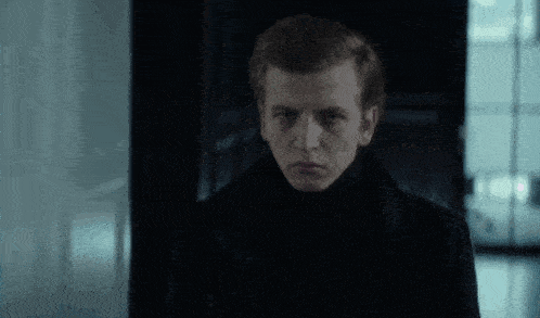 a young man in a black coat is standing in a dark room looking at the camera .