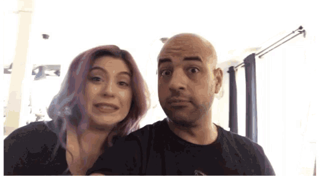 a man and a woman are posing for a picture together . the woman has pink hair and the man is bald .