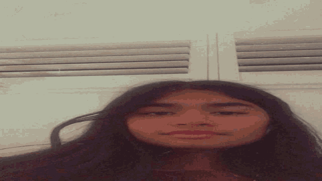 a girl with long dark hair is standing in front of a window with blinds