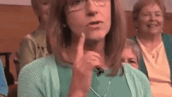 a woman wearing glasses and a green sweater is talking into a microphone while sitting in a crowd .