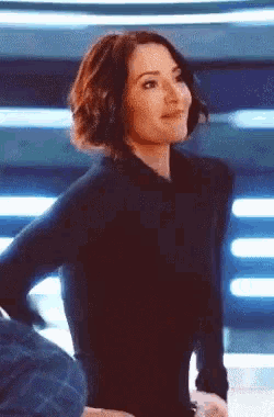 a woman in a black dress is standing with her hands behind her back and smiling .