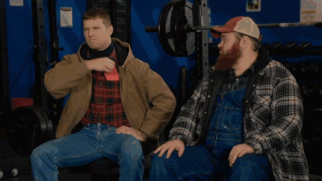 two men in plaid shirts and overalls sit on a bench in a gym