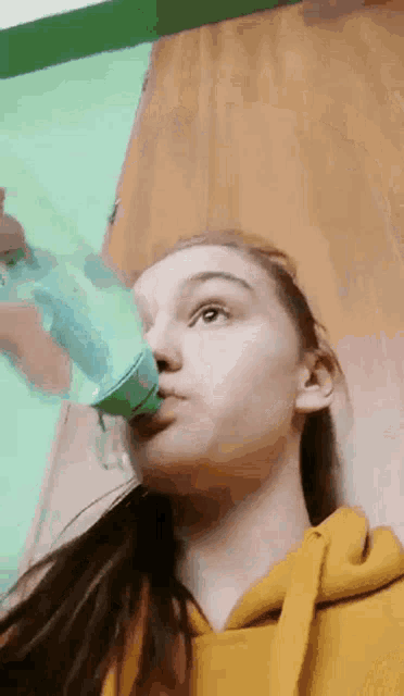 the girl is wearing a yellow hoodie and drinking water from a bottle .