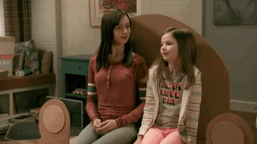 two girls are sitting on a cardboard chair in a living room . one girl is wearing a shirt that says happy love day .