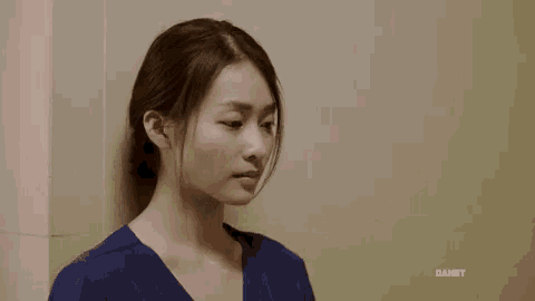 a woman in a blue scrub top is standing next to a wall and looking at the camera .