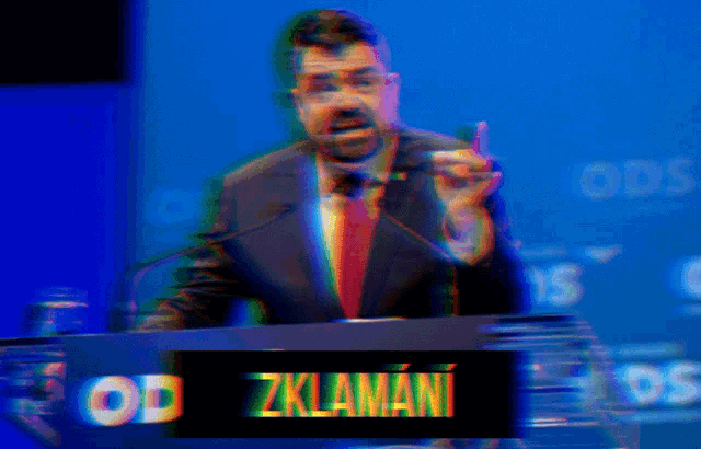 a man in a suit and tie stands at a podium with a sign that says zklamani