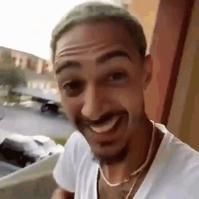 a man with a beard is smiling and wearing a white shirt and a gold chain around his neck .