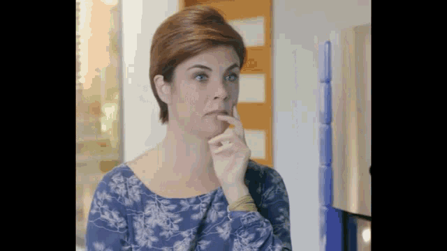 a woman in a blue floral shirt is holding her finger to her chin .