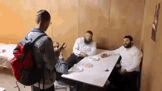 a man with a backpack is standing in front of a table with three men sitting at it .