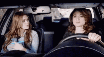 two women are sitting in a car talking to each other . one is driving and the other is sitting in the back seat .