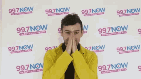 a man in a yellow jacket is standing in front of a wall with 99.7 now written on it