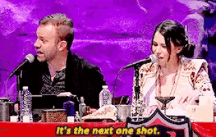 a man and a woman are sitting at a table talking into microphones with the words " it 's the next one shot " above them