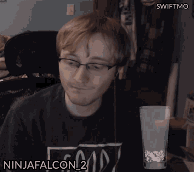 a man wearing glasses and a ninjafalcon 2 shirt looks at a glass of water