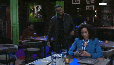 a man is standing next to a woman sitting at a table in a restaurant looking at her phone .
