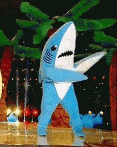 a blue and white shark mascot is standing in front of a water fountain