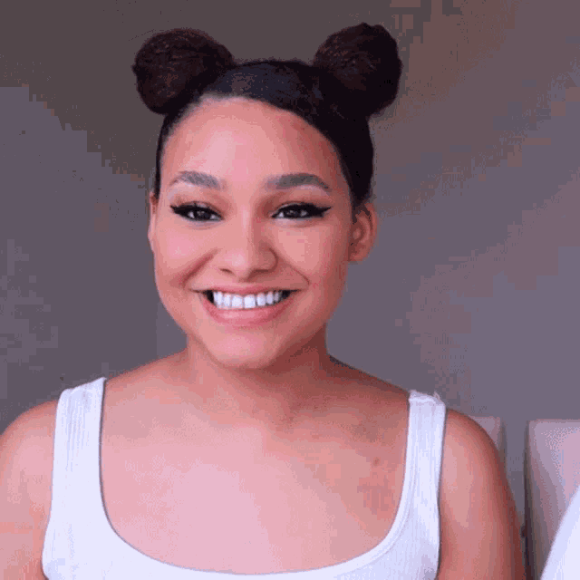 a woman wearing a white tank top with two buns in her hair smiles