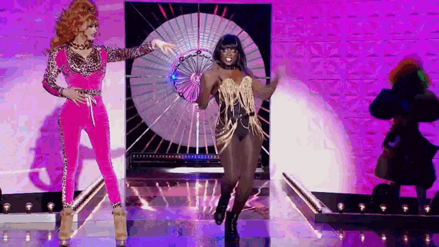 a couple of drag queens are dancing on a stage with a fan .