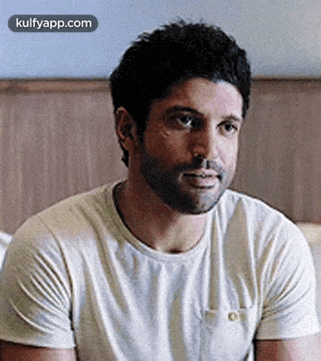 a man with a beard is sitting on a bed wearing a white shirt .
