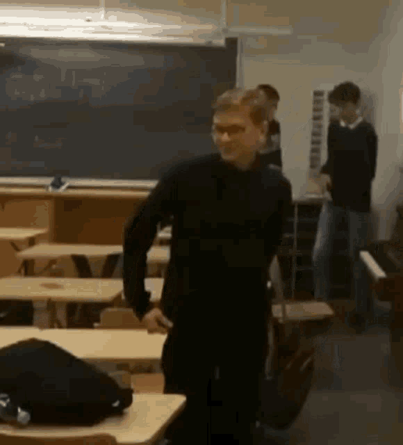 a man in a black shirt is standing in a classroom with a blackboard behind him