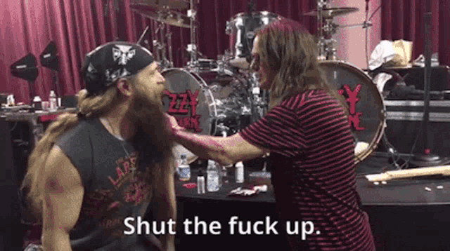 a man with a beard is being brushed by another man in front of a drum set with the words shut the fuck up below him