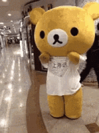 a yellow teddy bear mascot is wearing a white shirt and standing in a hallway .