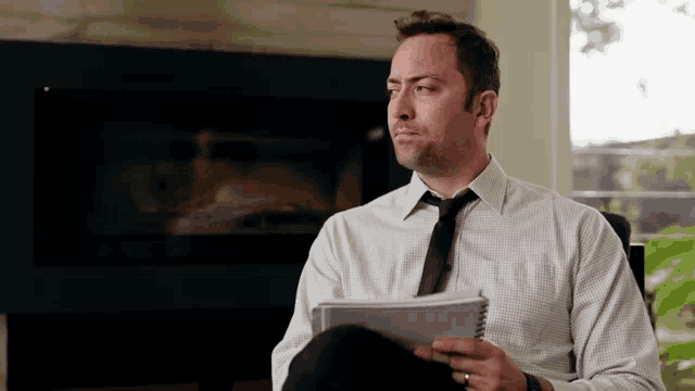 a man in a white shirt and tie is sitting in front of a fireplace and holding a notebook .