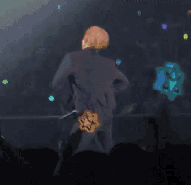 a man in a suit stands on a stage in front of a crowd with a glow in the dark sign that says m