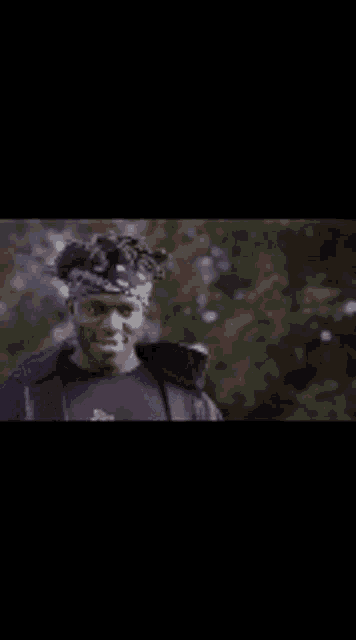 a man wearing a bandana and a black jacket is standing in front of trees .