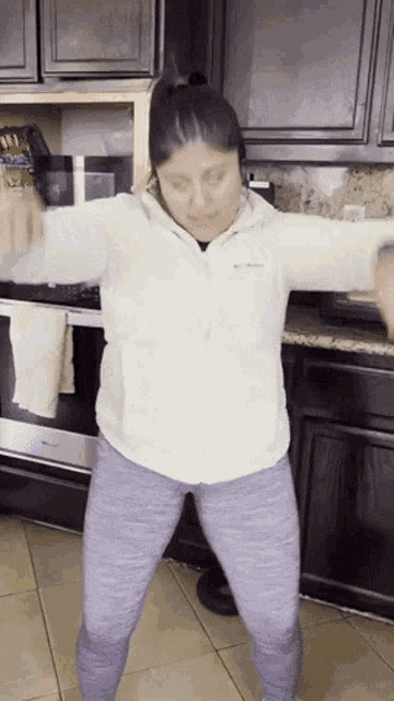 a woman wearing a white columbia jacket and purple leggings is dancing in a kitchen