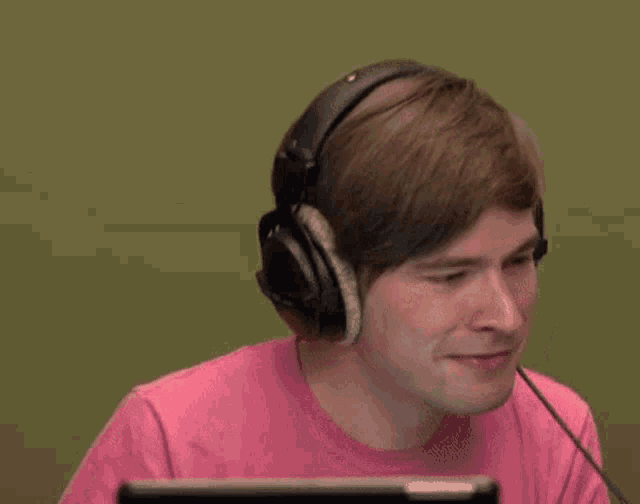 a man wearing headphones and a pink shirt is smiling while sitting in front of a computer .