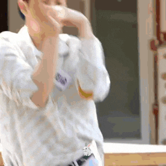 a man in a white shirt is covering his mouth with his hands while standing in front of a window .