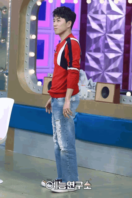 a man in a red sweater and ripped jeans is standing in front of a sign that says ' ae ' on it