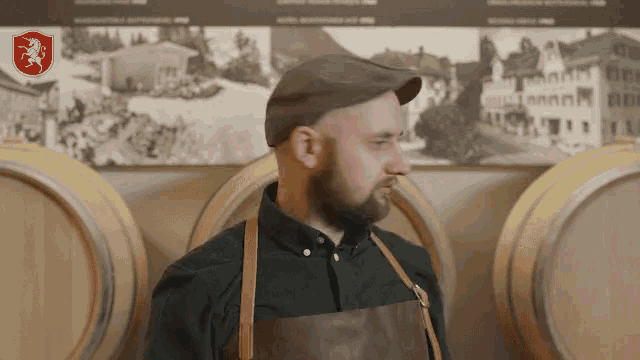 a man wearing a hat and apron stands in front of barrels with a picture of a horse on the wall behind him
