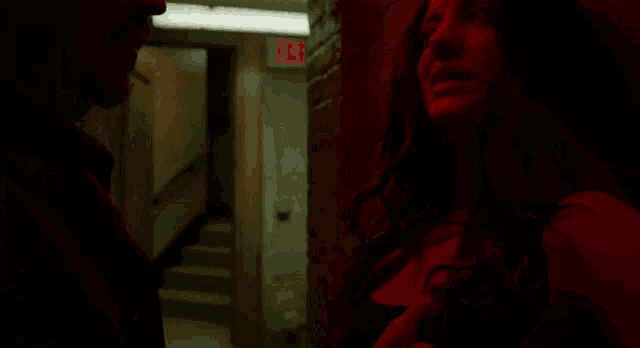 a man and a woman are standing in a dark room with a red exit sign in the background